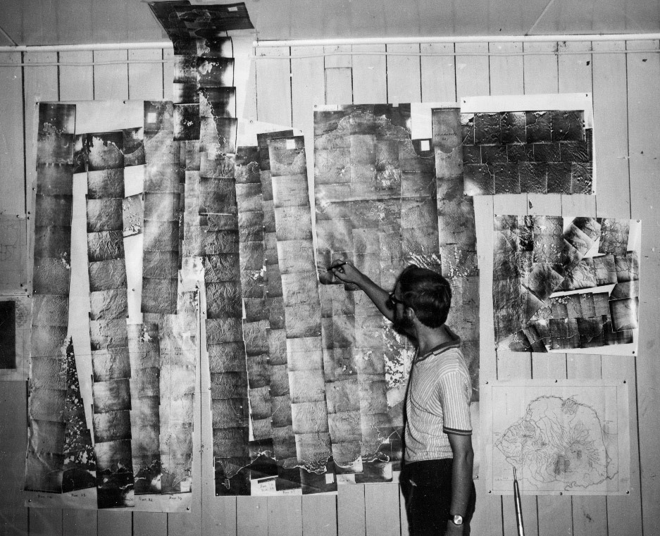 Black and white image showing a wall with long strips of photographs stuck to it. A man stands in front of the wall pointing at the images.