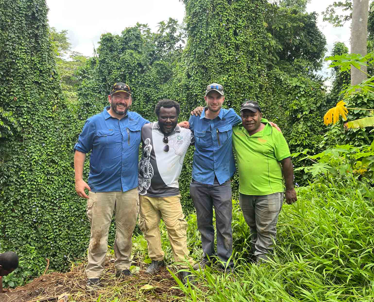Four men stand with arms around each other in a lush jungle landscape.