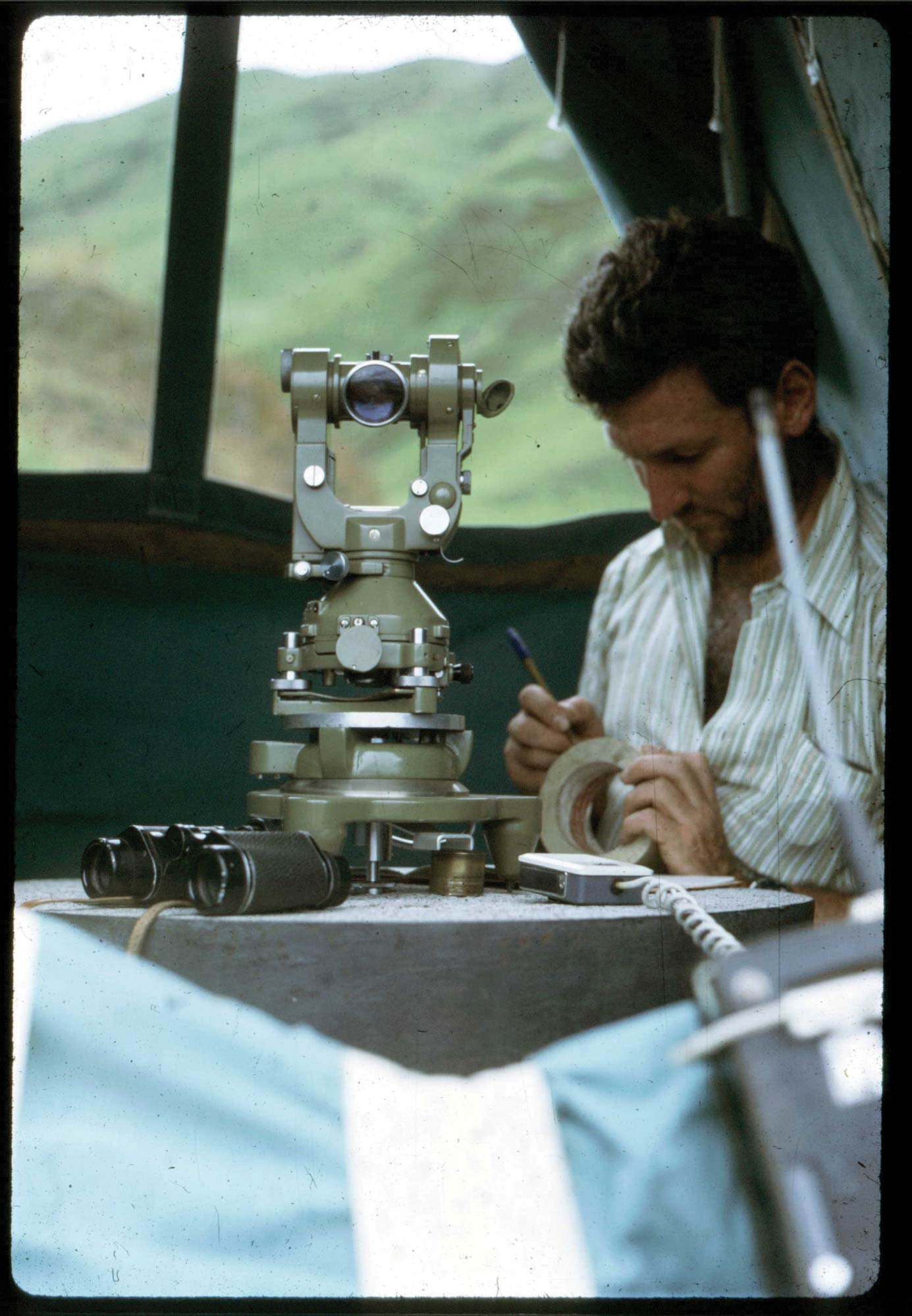 A man with a striped shirt and a beard sits at a table with a surveying instrument on it. He is making notes on a spool of tape.