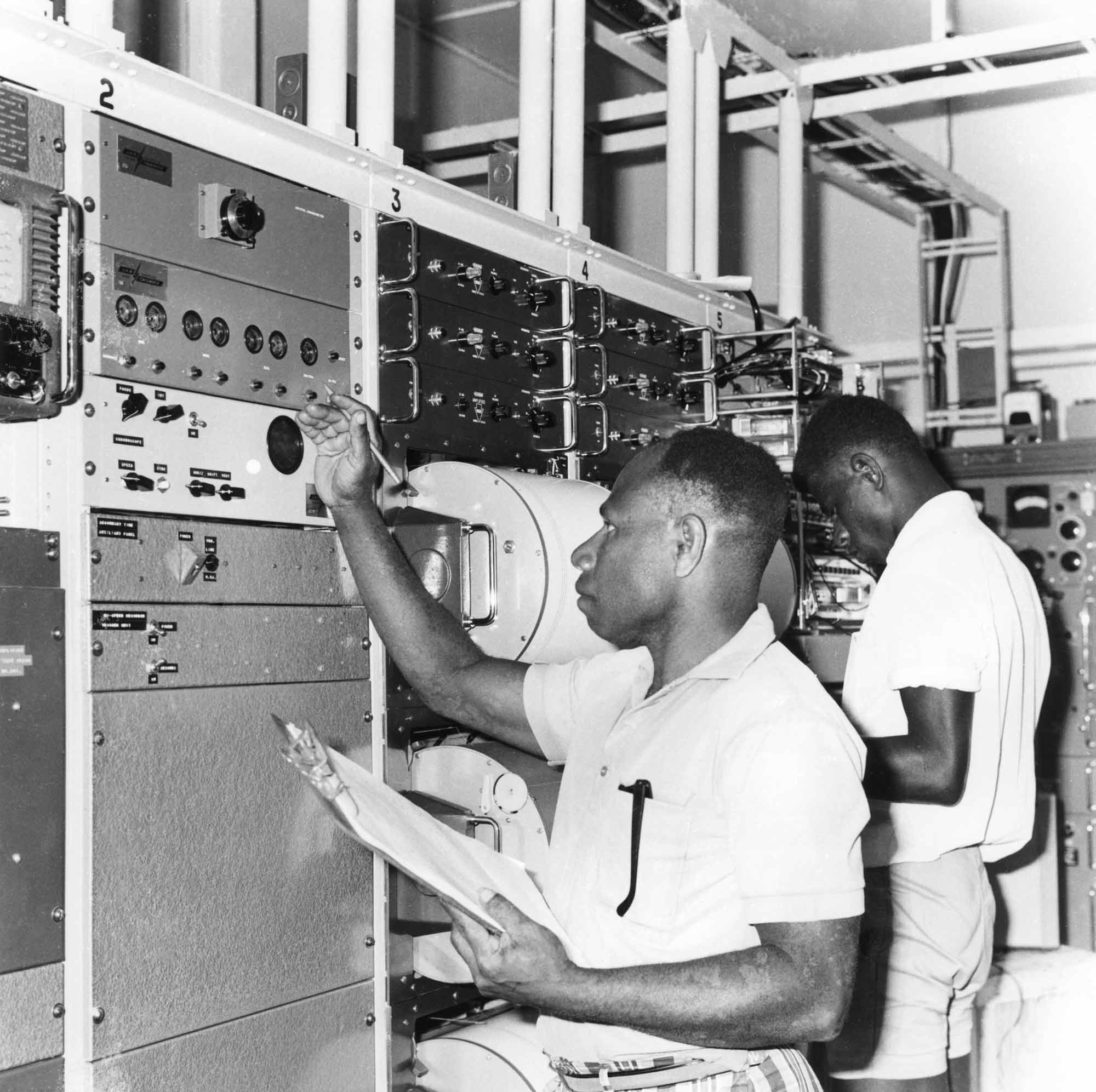 Black and white image of two men in white shirts working on a wall of complex scientific equipment.