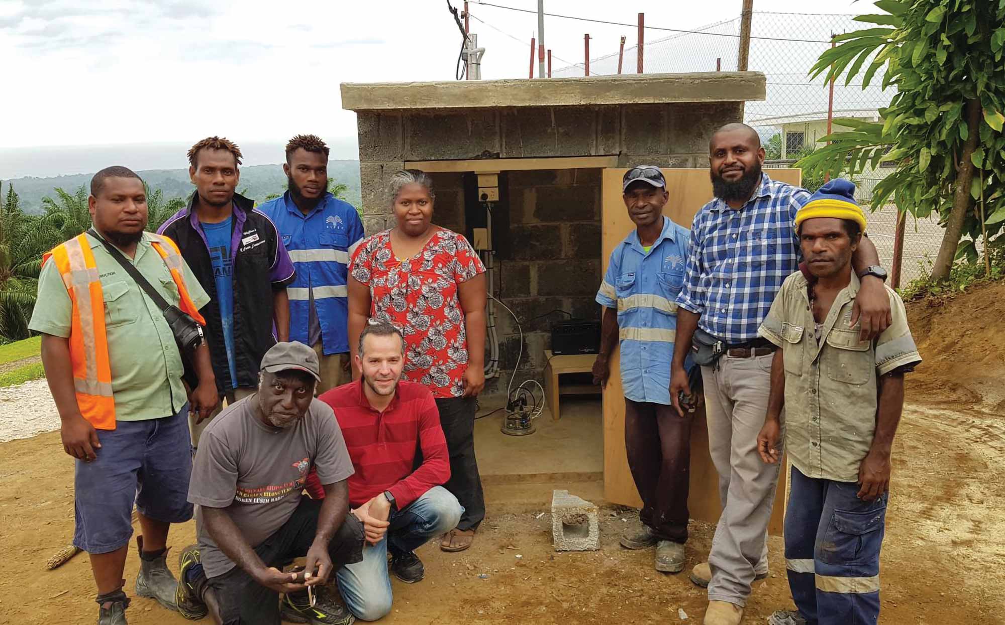 A group of nine men stand in front of a concrete hut that contains some scientific equipment.
