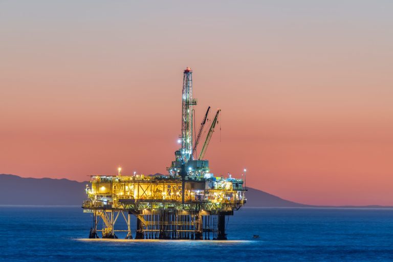 Dramatic image of an offshore oil platform off the coast against a pink winter sky as the sun sets and the rig's lights illuminate.