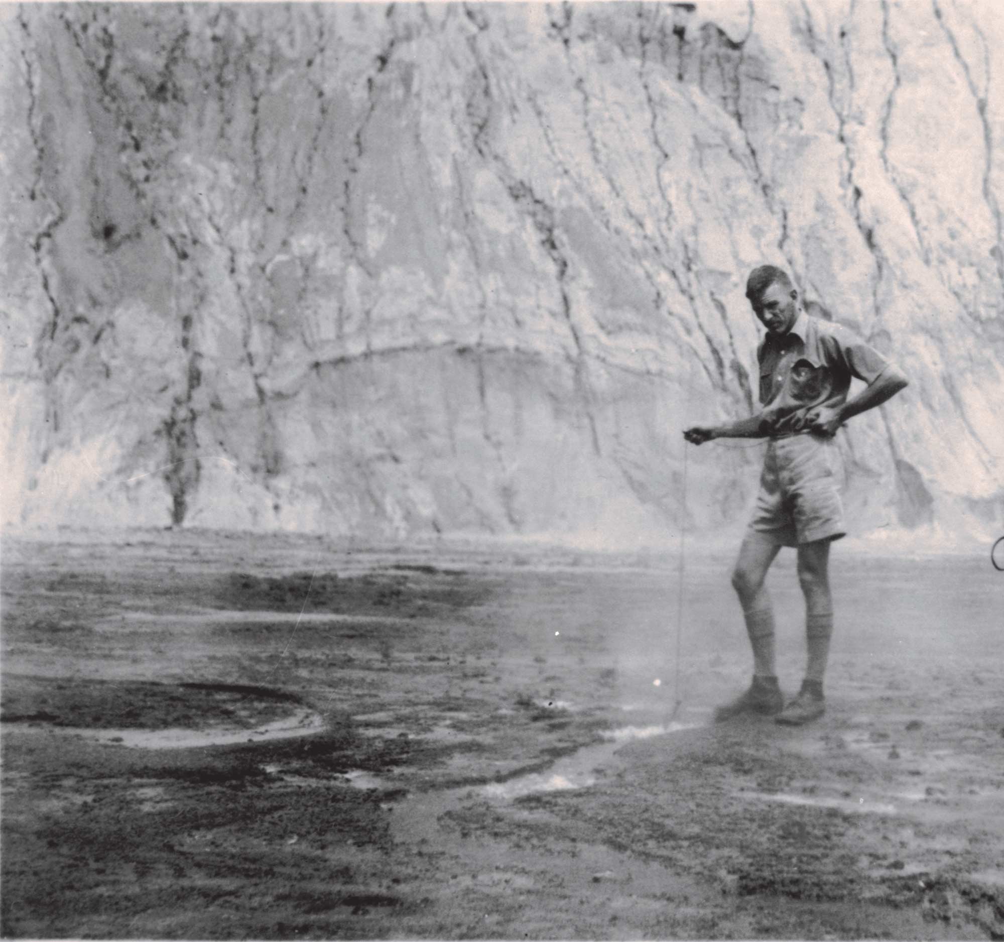 Black and white image of a man in shorts taking scientific measurements in a bare rocky landscape.