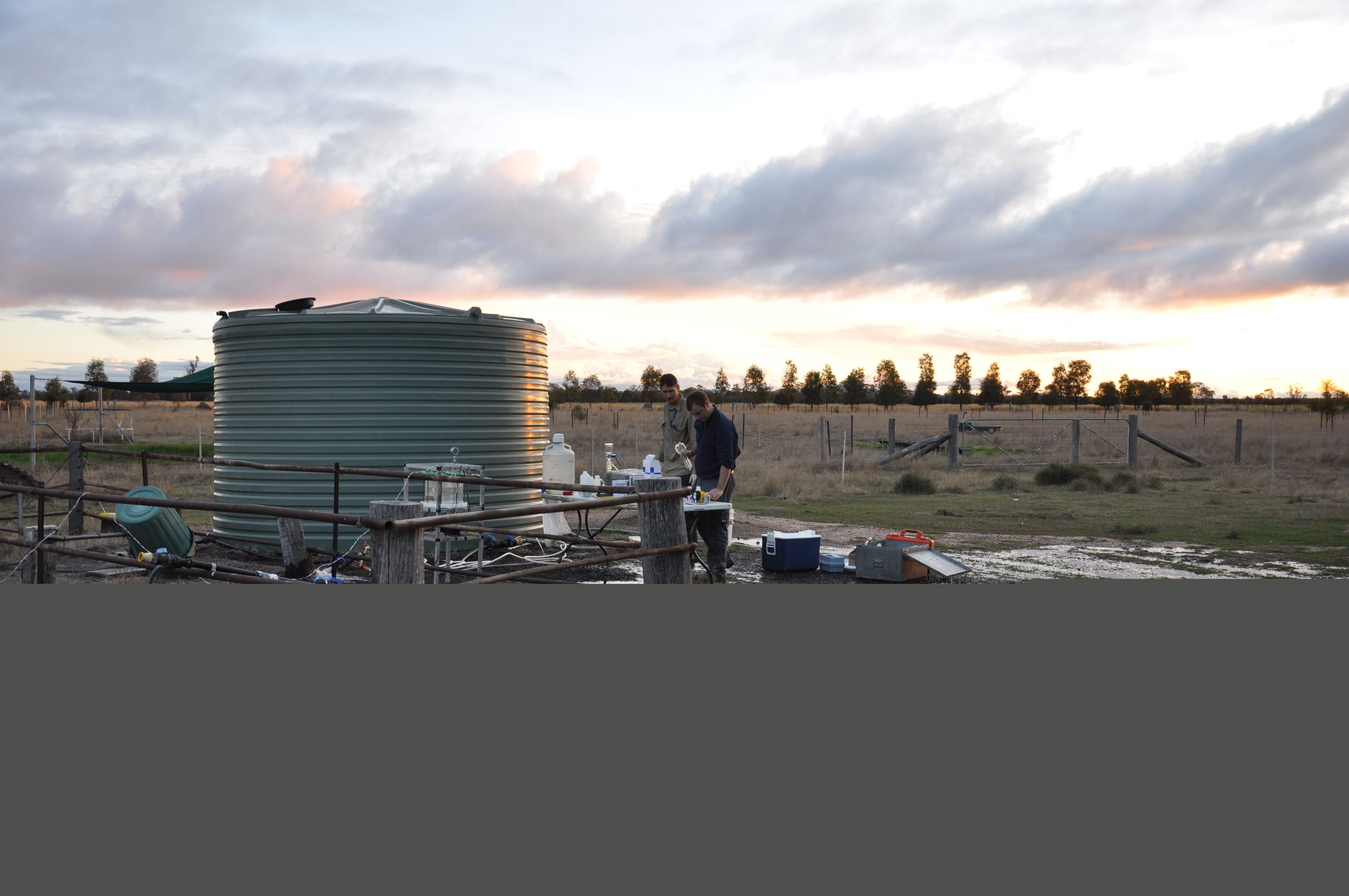 Two men standing near a paddock and a water tank with groundwater gas sampling equipment
