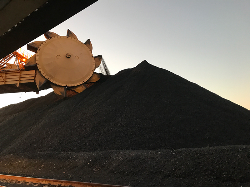 A pile of black coloured coal is being shifted by a large machine, called a bucket wheel excavator. A series of buckets are attached to a central wheel on the machine