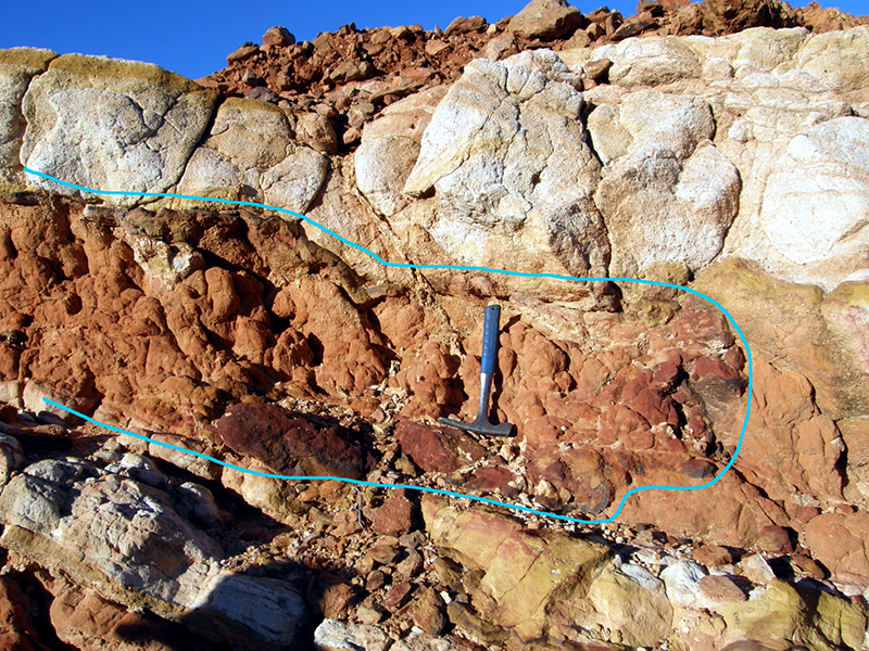A layer of orange roks sits between two layers of peler white and yeloow coloured rocks. A G-pick has been placed on a segment that is outlined by a blue line. This is the uranium roll front.