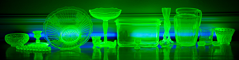 Various sized bowls, containers and drinking glasses glow green and sit on a dark table.