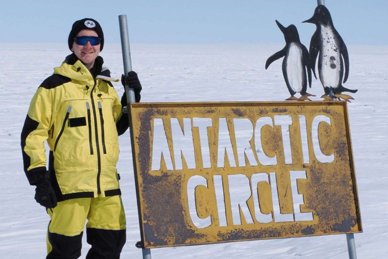 A man in bright yellow survival gear stands next to a metal sign that says 'Antarctic Circle'. All around is a flat icy landscape