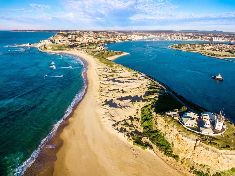 Image of A sandy isthmus and adjoining land in the distance, with blue ocean water running along either side of the isthmus.