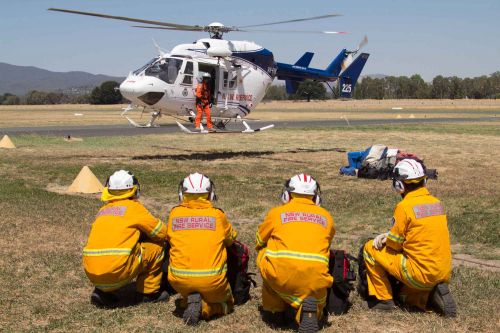 Four male firefighters crouch on ground with landed helicopter in distance